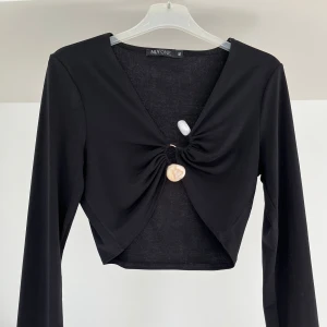Svart croppad topp med knyt från NLY ONE - Trendig svart croppad topp från NLY ONE med lång ärm och snygg knytning framtill. Toppen har en djup v-ringning och dekorativa detaljer vid knytet. Perfekt för att lyfta din outfit med en modern touch.
