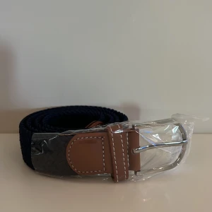 Mörkblått flätat bälte  - Snyggt mörkblått flätat bälte med bruna skinn-detaljer och silverfärgat spänne. Passar perfekt till jeans eller chinos och ger en stilren look. Bältet har vita sömmar på skinnet för extra detalj. 