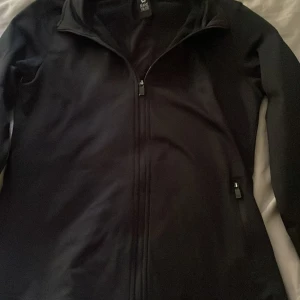 Svart träningströja från SOC med dragkedja - Säljer en svart träningströja från SOC i storlek 146-152. Tröjan har hel dragkedja framtill, hög krage och diskret logga på ryggen. Perfekt för dig som vill ha en enkel och stilren tröja till träning.