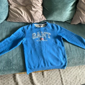 Blå sweatshirt från GANT - Säljer en blå sweatshirt från GANT med sydd logga och text på bröstet. Tröjan har rund halsringning och långa ärmar. Perfekt för en avslappnad stil och enkel att matcha med jeans eller joggers.