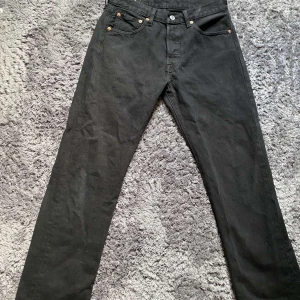 Svarta Levi's 501 jeans  - Klassiska svarta Levi's 501 jeans med raka ben och fem fickor. Jeansen har knappgylf, silverfärgade och kopparfärgade detaljer samt den ikoniska läderpatchen bak. Tillverkade i kraftigt jeanstyg som ger en tidlös look.