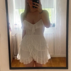 Vit volangklänning med smala axelband - Supersöt vit kortklänning med volanger och smala axelband. Klänningen har omlottkänsla och v-ringning, perfekt för sommarens alla tillfällen. Användes bara en gång