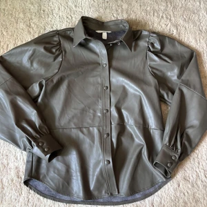 Mörkgrön jacka från H&M - Snygg mörkgrön jacka i skinnimitation från H&M med puffiga axlar och långa ärmar. Jackan har knappar framtill och vid ärmsluten, samt klassisk krage. Perfekt för dig som vill sticka ut lite extra.