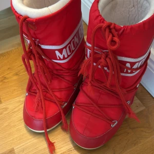 Röda Moon Boot snow boots - Säljer ett par klassiska röda Moon Boot snow boots med vit insida och tydlig logga på skaftet. Skorna har snörning framtill och är perfekta för vinterns kalla dagar. De har en rund tå och platt sula för extra komfort.