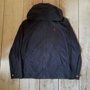 Marinblå vindjacka från Polo Ralph Lauren - Säljer nu min riktigt snygga marinblåa vindjacka från Polo Ralph Lauren med huva/utan. Har tagits hand om riktigt bra och den är äkthets kontrollerad! Riktigt snygg design och kan användas på soliga dagar och kallare och blåsigare dagar!