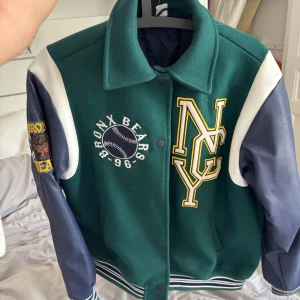 Grön bomberjacka  - Snygg grön bomberjacka från Divided med stora broderade patches, bland annat Bronx Bears och NY-logga. Jackan har blå och vita ärmar i skinnimitation, ribbade muddar och tryckknappar framtill. Oversized