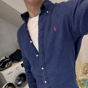 Blå Polo Ralph Lauren Linneskjorta - Snygg blå skjorta från Ralph Lauren i 100% linne. Storlek S. Klassisk modell med knappar hela vägen, button-down krage och den ikoniska röda polospelaren broderad på bröstet. Perfekt för sommaren.