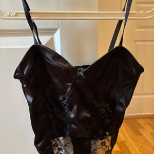 Svart korsett med spetsdetaljer - Snygg svart korsett med glansig finish och transparenta spetspaneler framtill. Justerbara axelband och liten rosett i mitten ger en extra touch. Perfekt för dig som vill ha en edgy och trendig look.