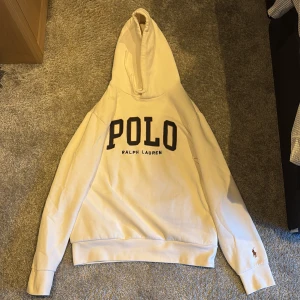 Vit hoodie från Polo Ralph Lauren - Storlek M. Nypris ca 1400. Säljer en klassisk vit hoodie från Polo Ralph Lauren med stor svart POLO-text och mindre RALPH LAUREN-tryck på bröstet. Tröjan har huva, långa ärmar och ribbade muddar. Liten broderad logga på ena ärmen. Perfekt för en avslappnad stil.