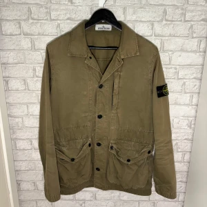Stone Island jacka - Grön overshirt jacka från Stone Island med klassisk krage och knappar framtill. Jackan har två stora fickor med lock och knapp. Perfekt för lager-på-lager-stil. Hör av dig vid intresse! 🙌