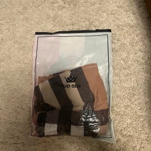 Beige och bruna rutiga shorts från burrbery - Säljer ett par snygga shorts från burberry med stora rutor i beige, brunt, svart och vitt samt en röd rand. Shortsen har resår i midjan med dragsko och en bakficka. Perfekta för sommaren!