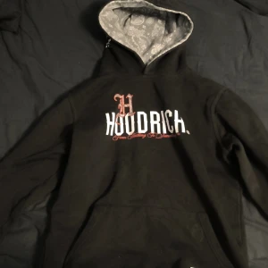 Svart Hoodie från Hoodrich XL junior storlek 13 -15 år - Svart hoodie från Hoodrich med stor vit och röd logga framtill och tryck på ryggen. Insidan av huvan har grått paisleymönster. Tillverkad i 80% bomull och 20% polyester. Klassisk känguruficka och huva för extra streetstyle. 