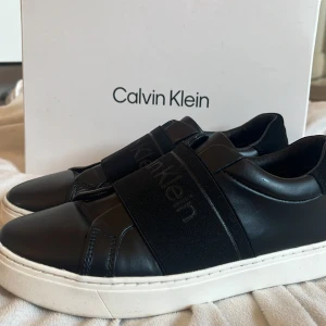 Svarta sneakers från Calvin Klein - Stilrena svarta sneakers från Calvin Klein med bred elastisk rem över foten där loggan syns diskret. Skorna har vit platt sula med CK-mönster undertill och är tillverkade i skinnliknande material. Perfekta för en clean och modern look.