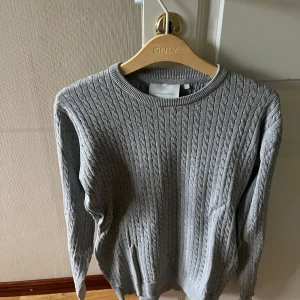 Grå kabelstickad tröja från Tailored Originals - Snygg grå kabelstickad tröja med rund halsringning och långa ärmar. Tröjan har ett klassiskt flätmönster och är perfekt för lager-på-lager-stil. Skön och stilren att matcha med jeans eller kjol.