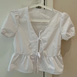Vit blus med puffärm och knyt - Söt vit blus med korta puffärmar, v-ringning och knyt framtill. Blusen har volang nertill och är tillverkad i bomull. Perfekt för en fräsch och somrig look. Ny 
