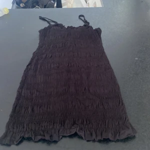 Svart smockad klänning med volang - Säljer en svart kortklänning med smockad struktur och volangdetaljer nertill. Klänningen har smala axelband och är perfekt för varma dagar. Enkel och stilren design som passar till många tillfällen.