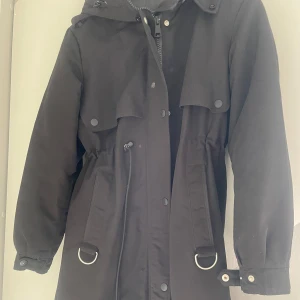Svart parkajacka från Copperose - Säljer en svart parkajacka från Copperose i storlek S. Jackan har huva, dragsko i midjan, knappar och dragkedja framtill samt praktiska fickor. Snygga detaljer med metallringar och justerbara ärmslut. Perfekt för kyligare dagar! Plus med äkta päls inköpspris 1900 kr 