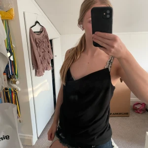 Svart linnetopp  - Svart linnetopp med tunna axelband. Toppen har en lös passform och ett mjukt, lite glansigt tyg som ger en stilren look. Perfekt för dig som gillar en enkel men ändå chic stil.