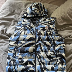Moncler dunväst med blå camomönster - Säljer en fet dunväst från Moncler med blå, svart och vit camouflage. Västen har huva, dragkedja framtill och två fickor med dragkedja. Ikonisk Moncler-logga på bröstet. Perfekt för dig som vill sticka ut med streetstyle.