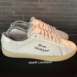 Vita Saint Laurent sneakers i canvas - Snygga vita sneakers från Saint Laurent med handskriven logga på sidan. Skorna har en klassisk låg profil, rund tå och platt sula i beige gummi. Ovandelen är i canvas med råa kanter och vita snören för en avslappnad vibe. TAR OCKSÅ HELST EMOT BETYDEN.