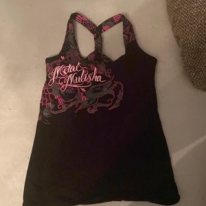 Metal Mulisha svart linne med tryck - Svart linne från Metal Mulisha med rosa och grått tryck framtill och coolt korsade axelband med mönster. Snyggt tryck med logga och detaljer i rosa och grått. Perfekt för dig som gillar streetwear och edgy stil.