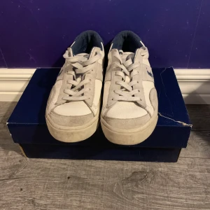 Vita sneakers från Polo Ralph Lauren. Man får med även lådan  - Snygga vita sneakers från Polo Ralph Lauren med klassisk blå logga på sidan. Skorna har rund tå, platt sula och är tillverkade i skinn och syntet med blå detaljer på insidan. Perfekta för en clean och sportig look.