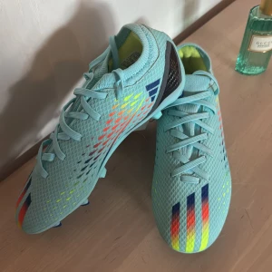 Adidas fotbollsskor - Speedportal  - Säljer ett par turkosa fotbollsskor från Adidas med färgglada ränder i blått, rött, gult och orange framtill. Skorna har snörning och mönstrad ovandel, samt dobbar under sulan för bra grepp på planen.