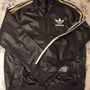 Svart Adidas track jacket - Säljer en svart Adidas track jacket med guldiga och vita ränder på axlarna. Jackan har dragkedja, ståkrage och stora Adidas-loggor både fram och bak. 