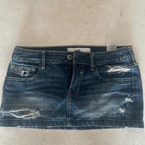 Jeanskjol från Hollister - Säljer en snygg blå jeanskjol från Hollister med slitna detaljer och broderad logga på fickan. Kjolen är kort och har klassisk femficksmodell med knapp och dragkedja framtill. Perfekt för en avslappnad och trendig look.