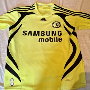 Chelsea borta 2007 gul med svarta detaljer - Chelsea FC borta 2007 tröja i gult med svarta mönster och Adidas-logga. Stort tryck 'Samsung Mobile' på bröstet och klubbmärke. Nummer 26 och namnet 'Terry' på ryggen. Klassisk design med korta ärmar och sportig känsla. Storlek 164. Tröjan är riktig kostar 1400 som ny