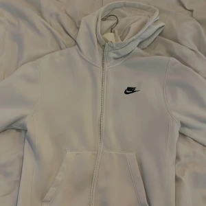 Vit hoodie med dragkedja från Nike - Säljer en stilren vit hoodie från Nike med dragkedja framtill och klassisk logga på bröstet. Tröjan har huva och två fickor framtill, perfekt för en sportig och avslappnad look.