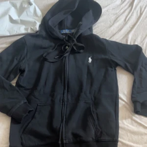 Svart hoodie från Polo Ralph Lauren - Svart hoodie från Polo Ralph Lauren med vit broderad logga på bröstet. Tröjan har dragkedja, huva med snörning och två fickor framtill. Custom fit-modell i mjuk bomull, perfekt för en chill och stilren look.