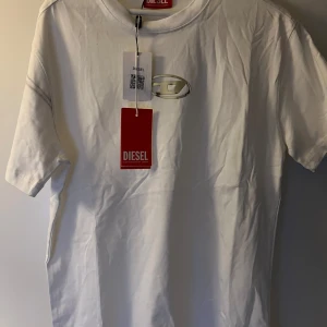 Vit t-shirt från Diesel med logga - Snygg vit t-shirt från Diesel med diskret logga framtill. Klassisk passform med korta ärmar och rund halsringning. Perfekt för dig som gillar stilrena och trendiga plagg. Materialet är mjuk bomull som känns skönt mot huden.