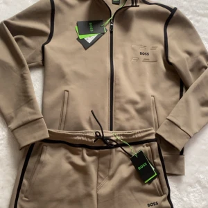 Beige mjukisbyxor från BOSS med svarta detaljer - Snygga beige mjukisbyxor från BOSS med svarta kontrastlinjer längs sidorna och logga på benet. Byxorna har dragsko i midjan och är tillverkade i mjukt material för en bekväm känsla. Perfekta för en avslappnad stil. Säljs pga fel storlek (fick i present) 