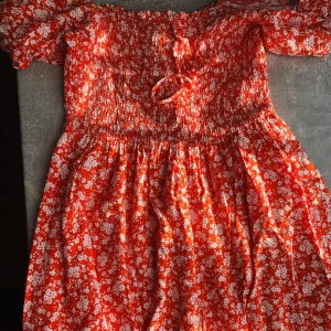 Röd blommig offshoulder klänning - Söt röd klänning med vita blommor och offshoulder-modell. Smockad överdel med knyt framtill och korta ärmar. Perfekt för varma dagar och somriga tillfällen.