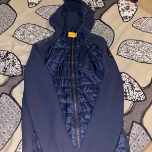 Blå Parajumpers pufferjacka med huva - Snygg marinblå pufferjacka från Parajumpers med huva och dragkedja. Jackan har quiltade partier framtill och baktill, samt mjuka ärmar och sidor i polyester. Praktisk bröstficka med dragkedja och cool gul detalj vid halsen. Perfekt för kyliga dagar.