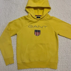 Gul GANT hoodie  - Gul hoodie från GANT med broderad logga på bröstet. Ficka framtill, huva och långa ärmar. Mjuk bomullskvalitet och använd ett fåtal gånger.  Säljs pga för liten.