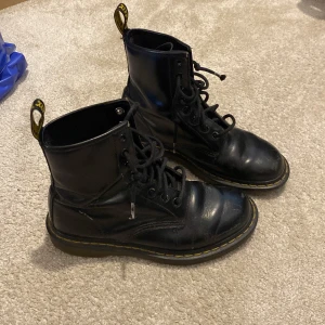 Svarta Dr. Martens boots i skinn - Klassiska svarta Dr. Martens boots i skinn med rund tå, snörning och den ikoniska gula sömmen runt sulan. Grov platt sula och dragflik baktill med gul text. Synligt använda med lite repor och creases (syns på bild).