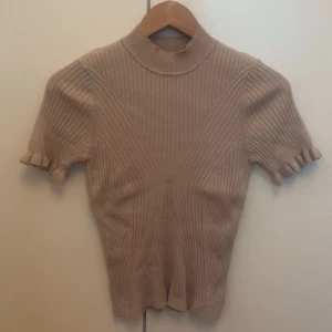 stickad topp med volangärm - Superfin beige stickad topp med ribbat mönster och korta ärmar med volangkant. Högre halsringning och figurnära passform. Väldigt stretchigt material. Knappt använd