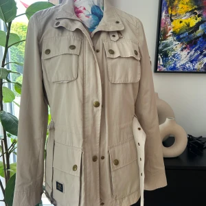 Beige jacka från Morris med fickor och knappar - Snygg beige jacka från Morris med fyra stora fickor framtill och dekorativa knappar. Jackan har hög krage, dragkedja och tryckknappar samt patch på ena ärmen. Nyligen kemtvättad. Perfekt för vår och höst.
