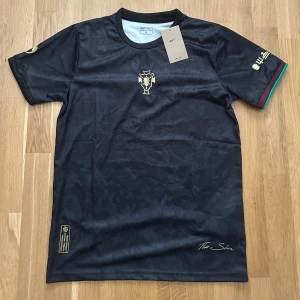 Svart Ronaldo Nike fotbollströja  - Svart fotbollströja från Nike i samarbete med The Sin och Common Goal. Guldiga broderade detaljer på bröstet och ärmen, samt diskret mönster i tyget. Ribbade muddar i vinrött och grönt på ena ärmen. Tillverkad i lätt och ventilerande material.