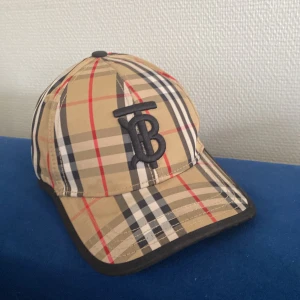 Burberry rutig keps med logga - Snygg keps från Burberry i klassiskt beige, svart, vit och röd rutigt mönster. Stor broderad TB-logga framtill och justerbart spänne bak med gulddetalj. Bak finns en patch med texten 'Burberry London England'. Insidan har Burberry-tryck längs sömmarna.