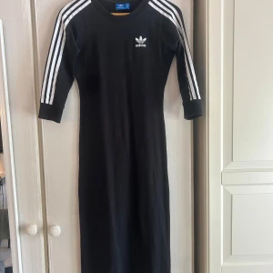 Svart Adidas klänning med ränder - Sportig svart klänning från Adidas med klassiska vita ränder längs ärmarna och broderad logga på bröstet. Modellen är lång, har trekvartsärm och är gjord i mjuk bomull. 