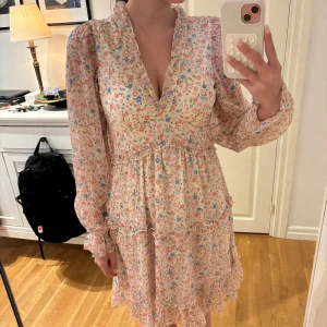 Klänning - Supersöt blommig klänning från Vero Moda i ljusa pastellfärger i storlek XS. Klänningen har volangdetaljer, v-ringning och långa ärmar. Perfekt för vår och sommar