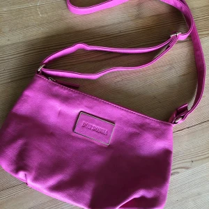 Rosa axelväska från Don Donna - Snygg retro rosa axelväska från Don Donna i syntetmaterial. Väskan har justerbar axelrem, dragkedja upptill och extra fack med dragkedja både på utsidan och insidan. Perfekt storlek för det viktigaste och ger en poppig färg till din outfit.
