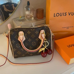 Louis Vuitton Speedy Bandoulière 20 Monogram - Säljer en klassisk Louis Vuitton Speedy Bandoulière 20 i monogram canvas med beige handtag och axelrem. Väskan har guldfärgade detaljer, dragkedja och ett körsbärs-berlock. Perfekt storlek för det viktigaste och ikoniskt LV-mönster.