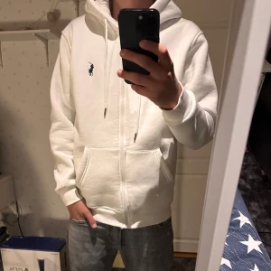 Vit hoodie från Polo Ralph Lauren - Säljer en klassisk vit hoodie från Polo Ralph Lauren med dragkedja och huva. Tröjan har broderad logga på bröstet, fickor framtill och ribbade muddar. Perfekt för en clean och stilren look.