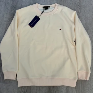 Vit sweatshirt från Tommy Hilfiger - Säljer en helt ny stilren vit sweatshirt från Tommy Hilfiger med klassisk rund halsringning och diskret broderad logga på bröstet. Tröjan har långa ärmar och ribbade muddar vid ärmslut och nederkant. Perfekt för en clean och avslappnad look.