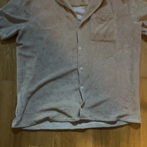 Beige frottéskjorta med knappar - Säljer en beige skjorta i mjuk frotté med kort ärm och klassisk krage. Skjortan har knäppning framtill och en bröstficka. Perfekt för en avslappnad stil.