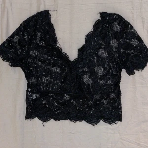 Svart spetsig croppad topp från SHEIN - Säljer en svart croppad topp från SHEIN med v-ringning och korta ärmar. Toppen är helt i spets med blommigt mönster och har fina vågiga kanter nertill! 💞